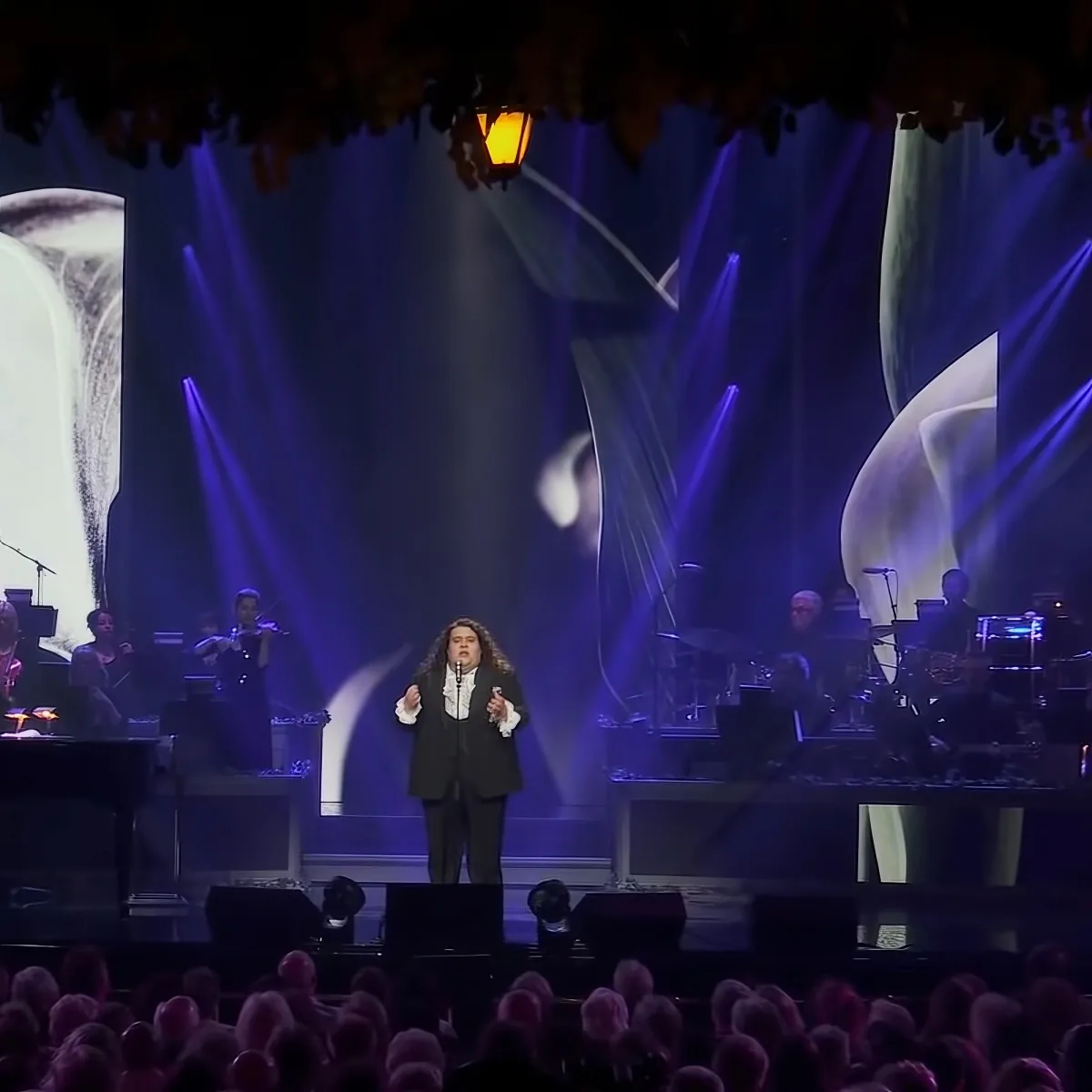 A person in formal attire sings on stage with a band and dramatic lighting.