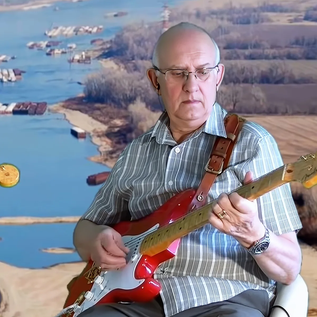 A man plays a red electric guitar, wearing a striped shirt and glasses, with a scenic background.