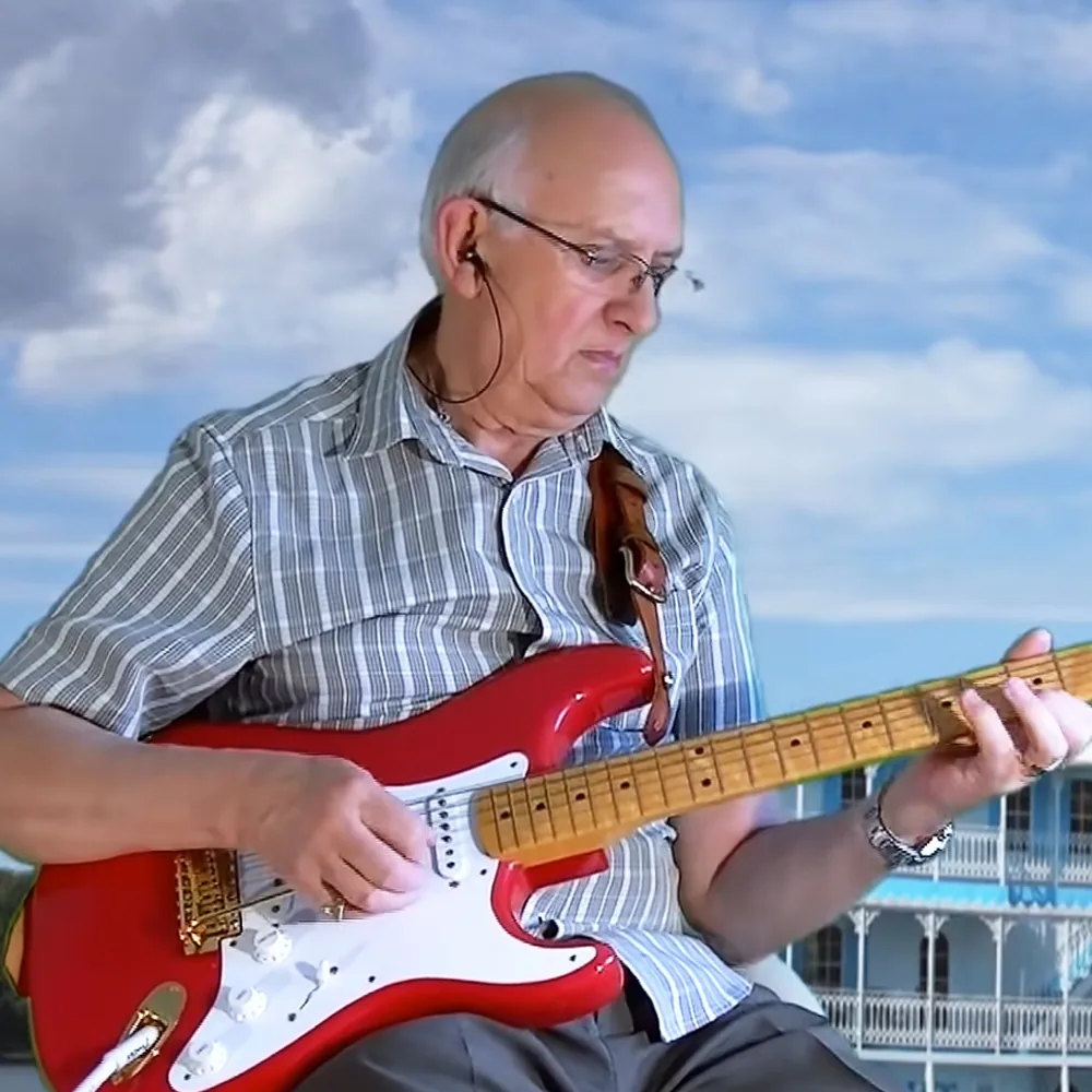 An older man plays a red electric guitar against a sky backdrop.