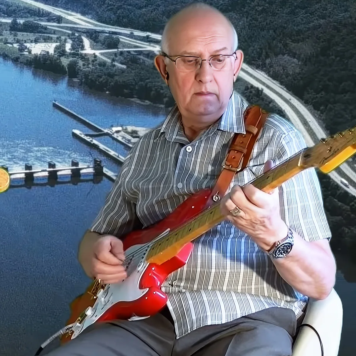 An older man plays a red electric guitar, wearing a striped shirt and glasses, with a scenic background of a river and...
