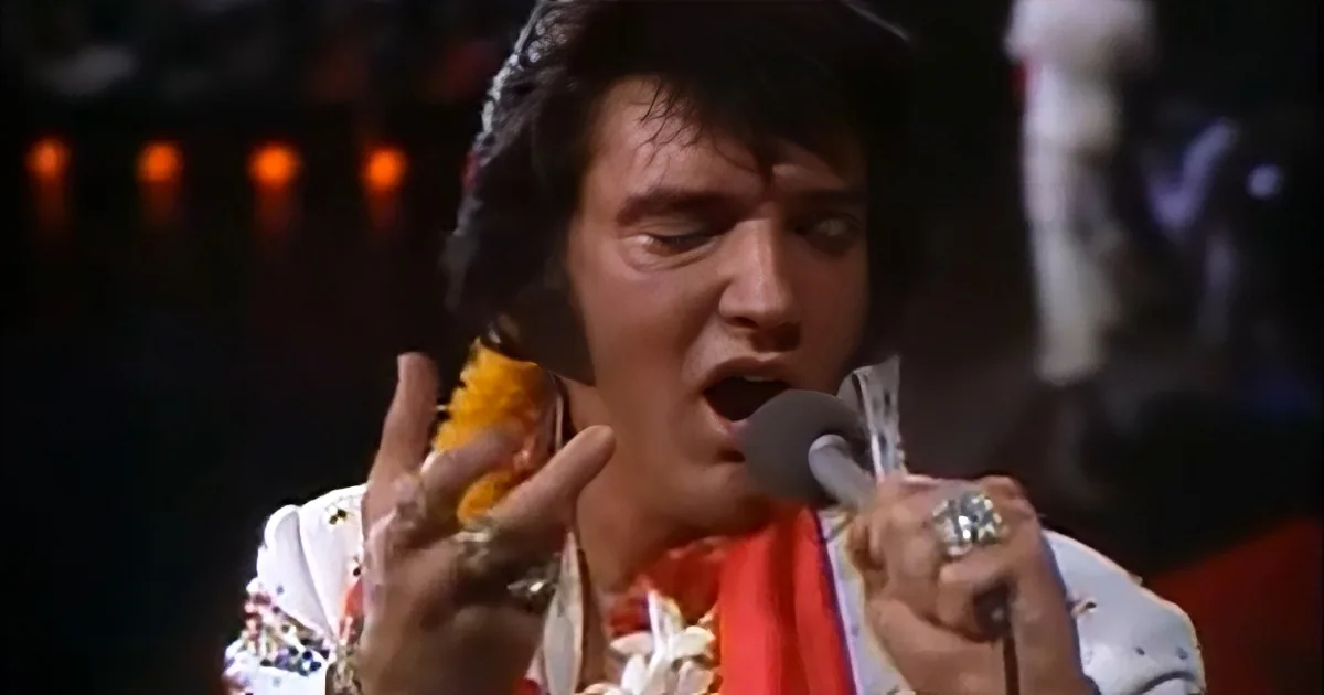 Elvis Presley in a white suit with a microphone, singing passionately on stage.