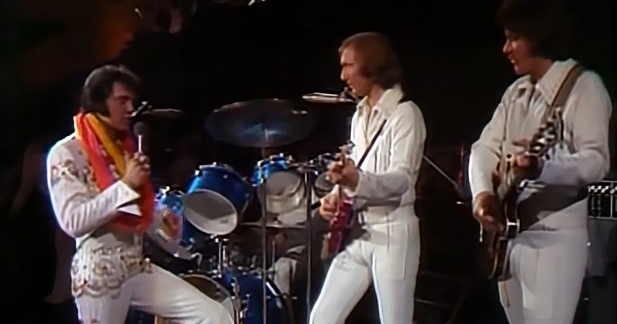Elvis Presley in white suit sings on stage with band, including saxophonist and drummer.