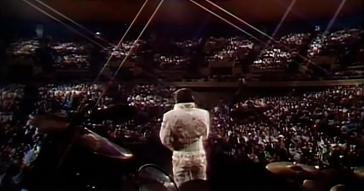 A person in a white outfit stands on stage facing a large, illuminated crowd.