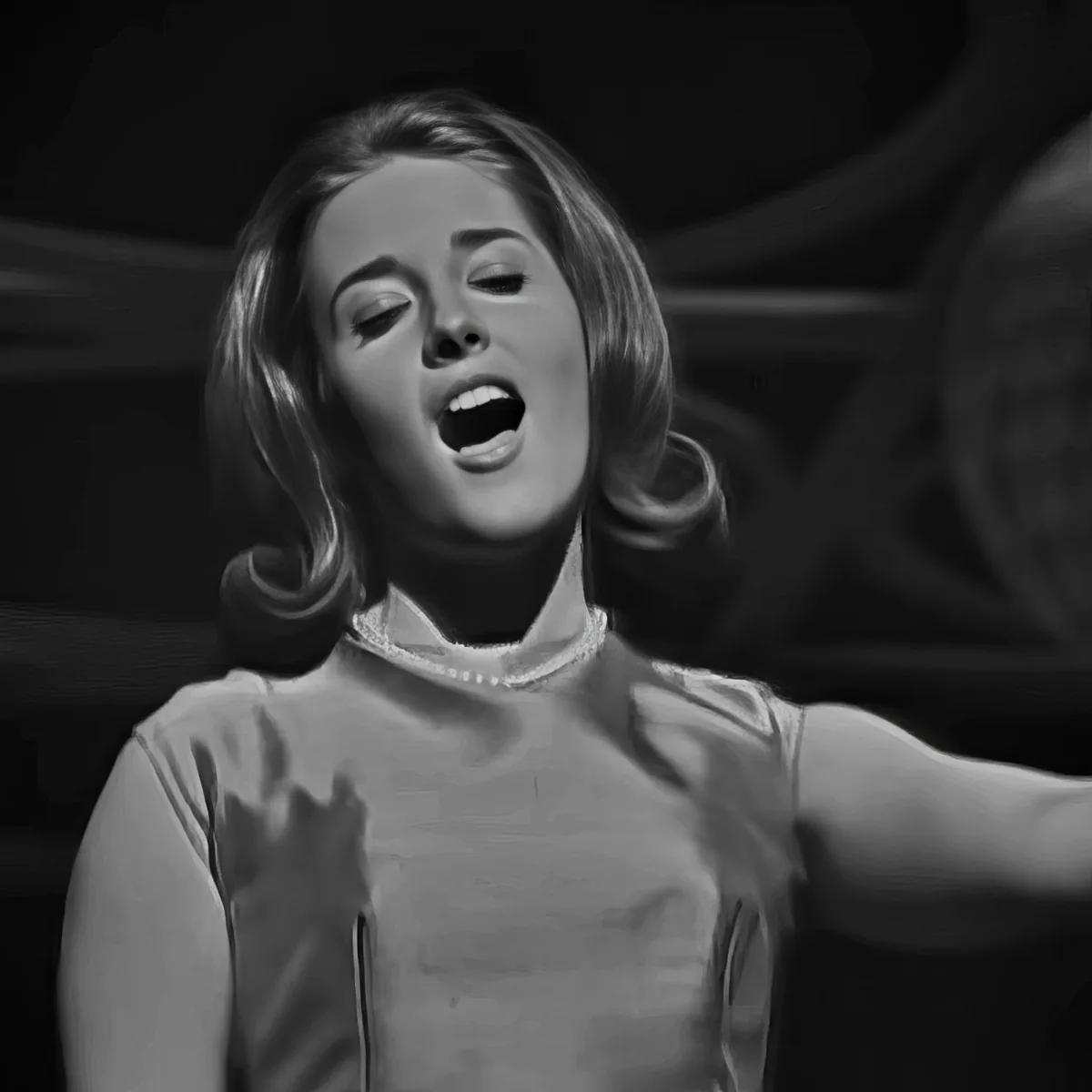A woman with shoulder-length hair sings passionately, wearing a light-colored top, in a black-and-white image.