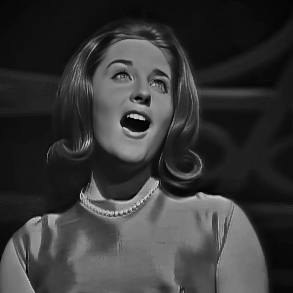 A woman with shoulder-length hair, wearing a pearl necklace, sings passionately in a black-and-white setting.