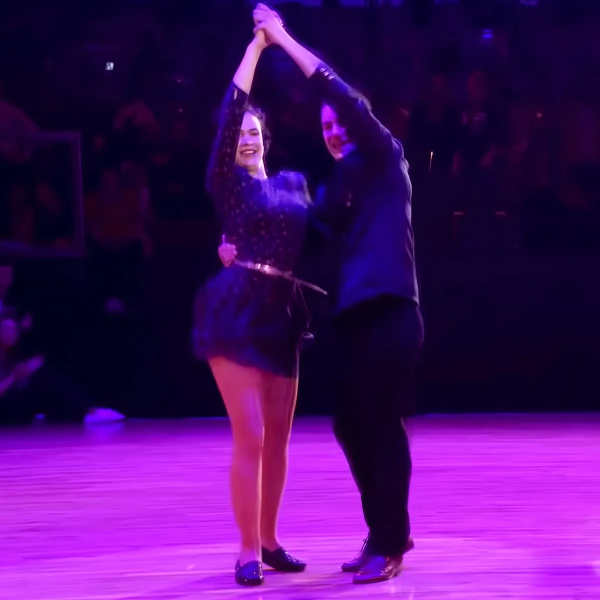 A couple in formal attire dances gracefully on a stage under purple lighting.