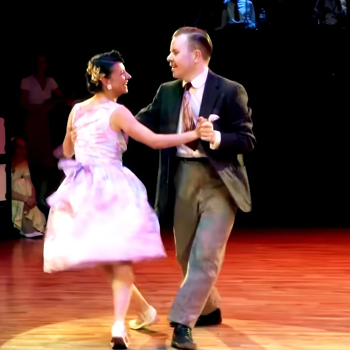 A couple in formal attire dances joyfully on a wooden floor, surrounded by other dancers.