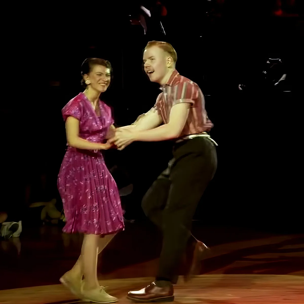 A man and woman are dancing together on stage, both smiling and holding hands.