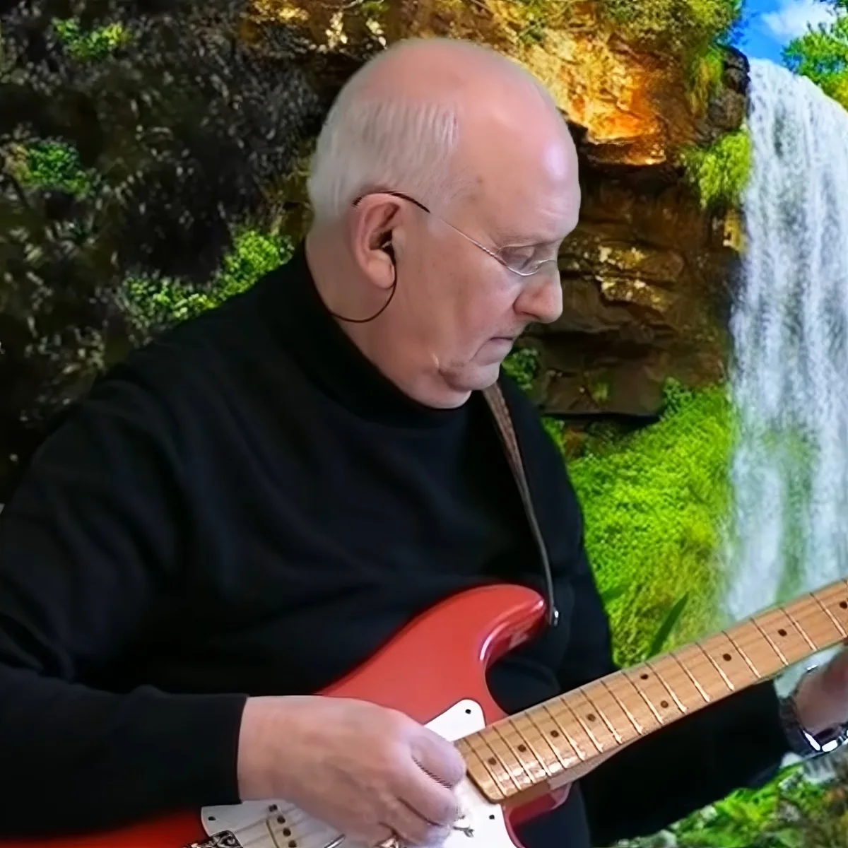 A man in a black shirt plays a red electric guitar with a waterfall and greenery in the background.