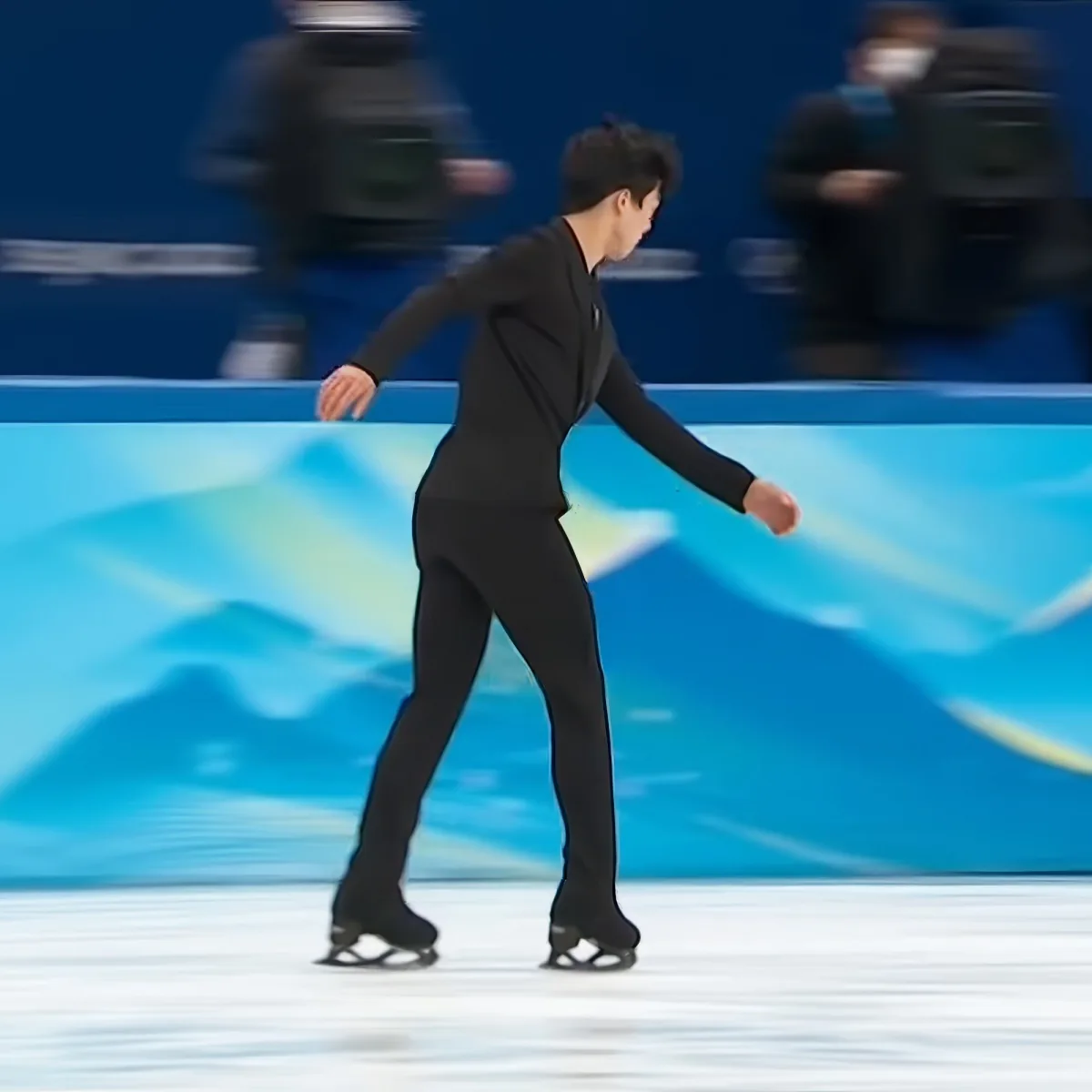 A figure skater in black attire glides on ice, arms outstretched, with blurred spectators in the background.