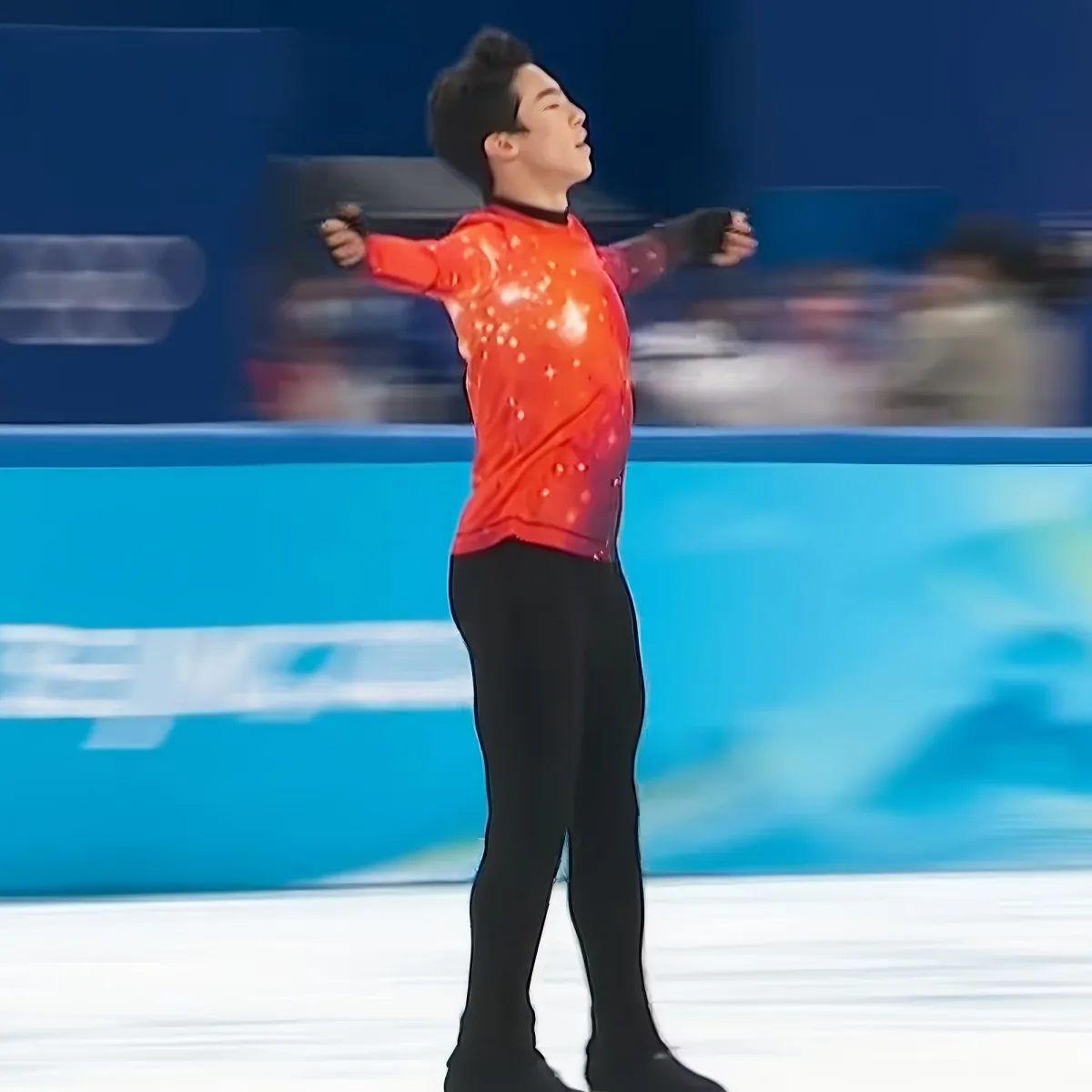 A figure skater in a red outfit with black pants stands on ice, arms outstretched, in a performance pose.