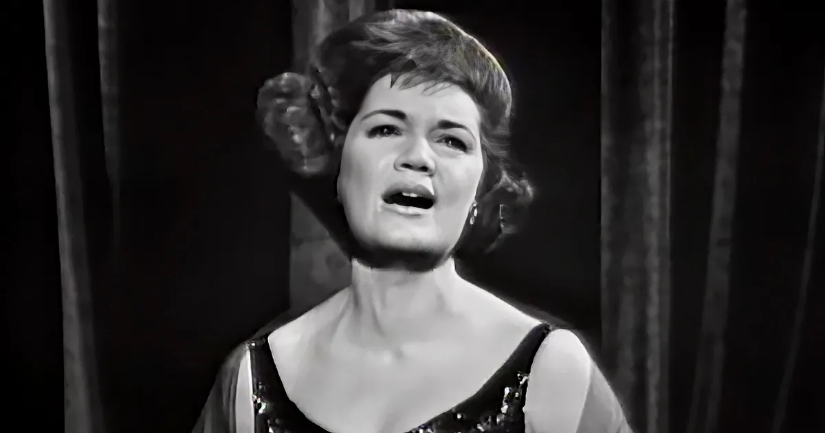 A woman in a black dress with a dramatic hairstyle and dark curtains in the background.