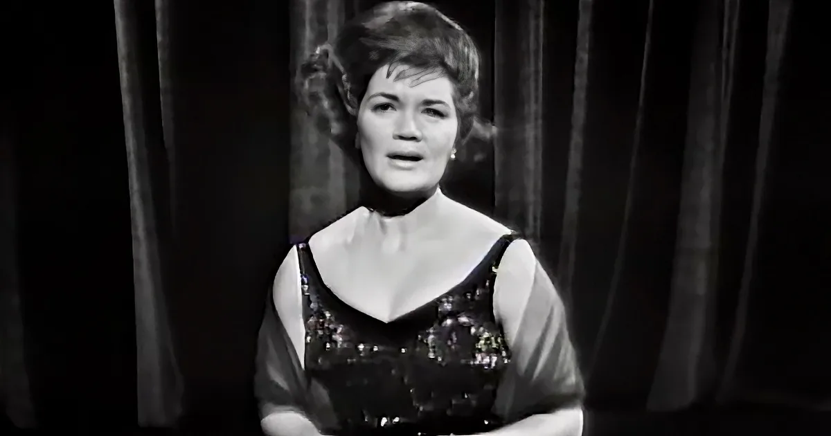A woman in a black dress with a floral pattern stands in front of dark curtains.
