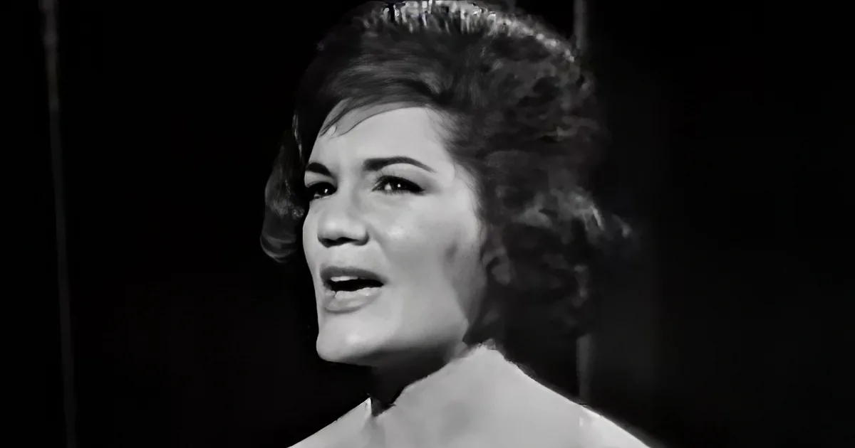 A woman with curly hair, wearing a white top, is singing with her mouth open.
