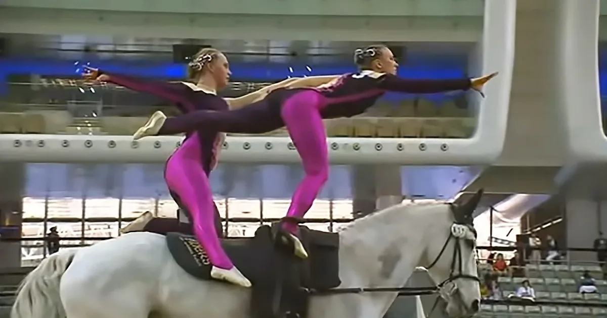 Two gymnasts in pink and black leotards perform a synchronized pose on a white horse in an indoor arena.