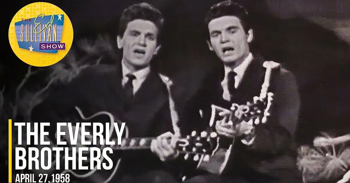 Two men in suits sing and play guitars, one with a floral pattern, against a dark background.