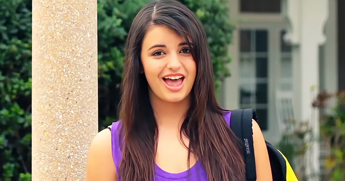 A smiling girl with long brown hair, wearing a purple top and carrying a backpack, stands outdoors near a pillar and...