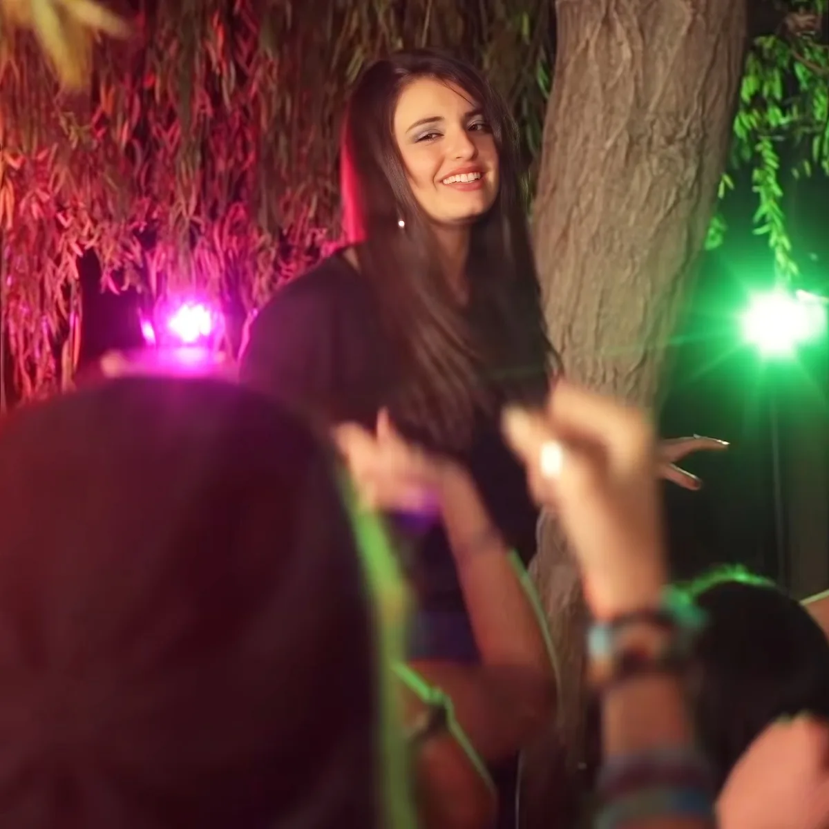 A woman with long hair smiles while holding a green string, surrounded by colorful lights and a tree.