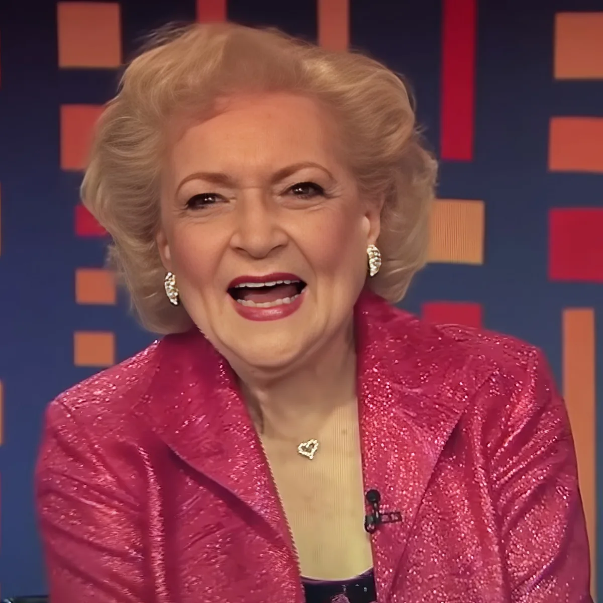 A woman with blonde hair, wearing a pink sequined jacket and heart-shaped necklace, smiles brightly on a colorful background.
