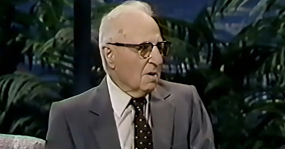 An older man in a suit and tie, wearing glasses, sits in front of a backdrop of green foliage.