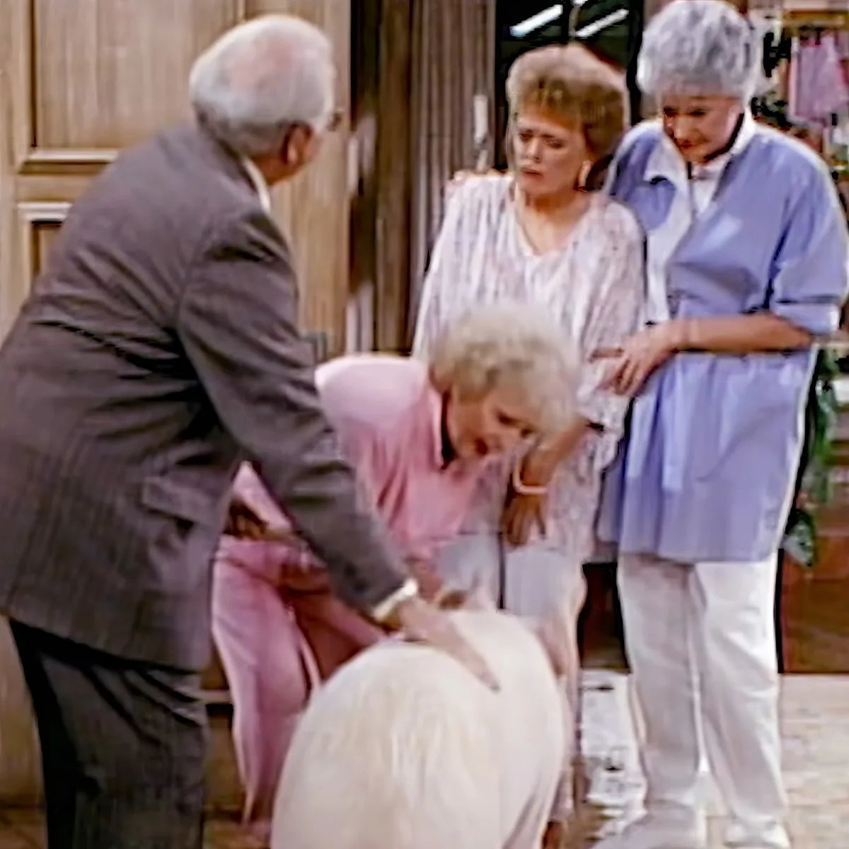 An elderly man in a suit leans over a woman in a pink dress, while others observe in a room.