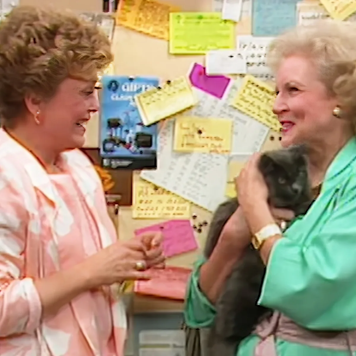 Two women, one holding a cat, smile and interact in a room with a bulletin board full of notes.