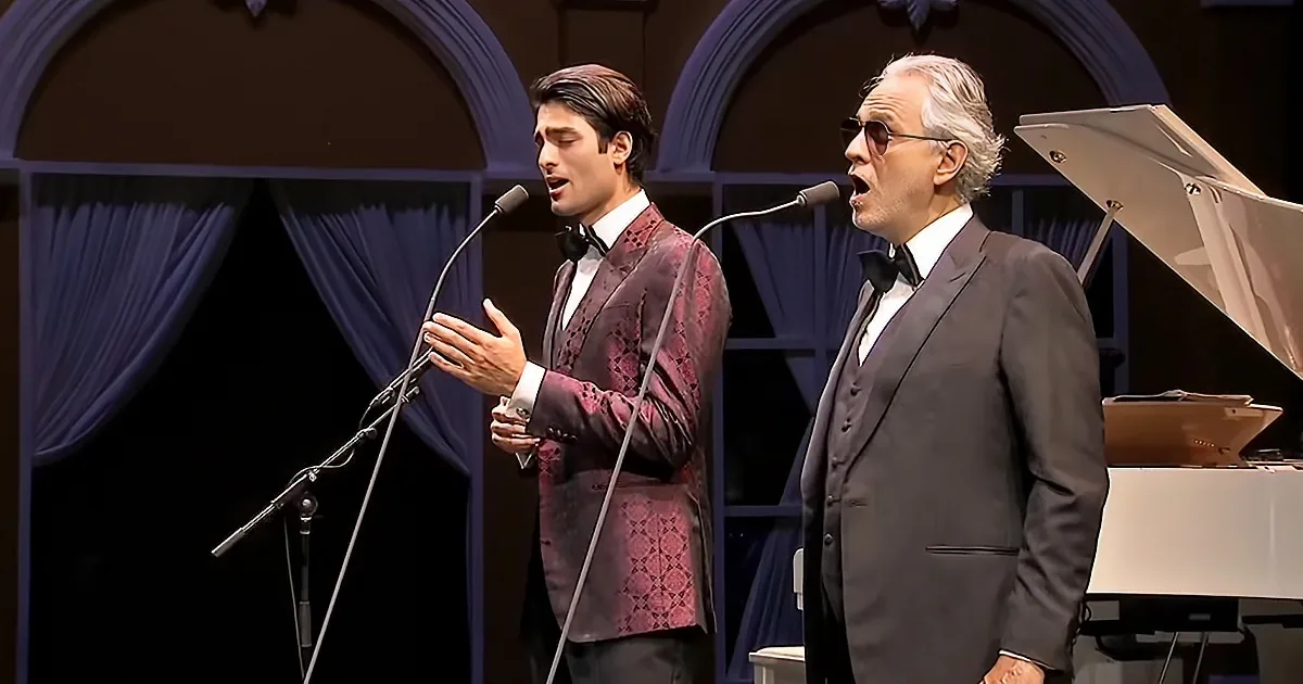 Two men in suits sing into microphones on stage with a piano in the background.