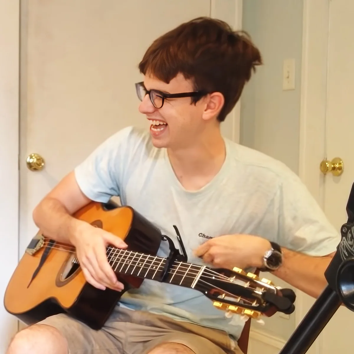A person wearing glasses and a light shirt is playing a guitar while smiling.