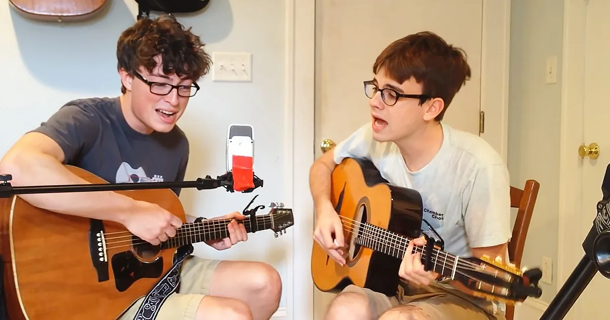 Two young men are playing acoustic guitars and singing in a room with microphones and a phone setup.