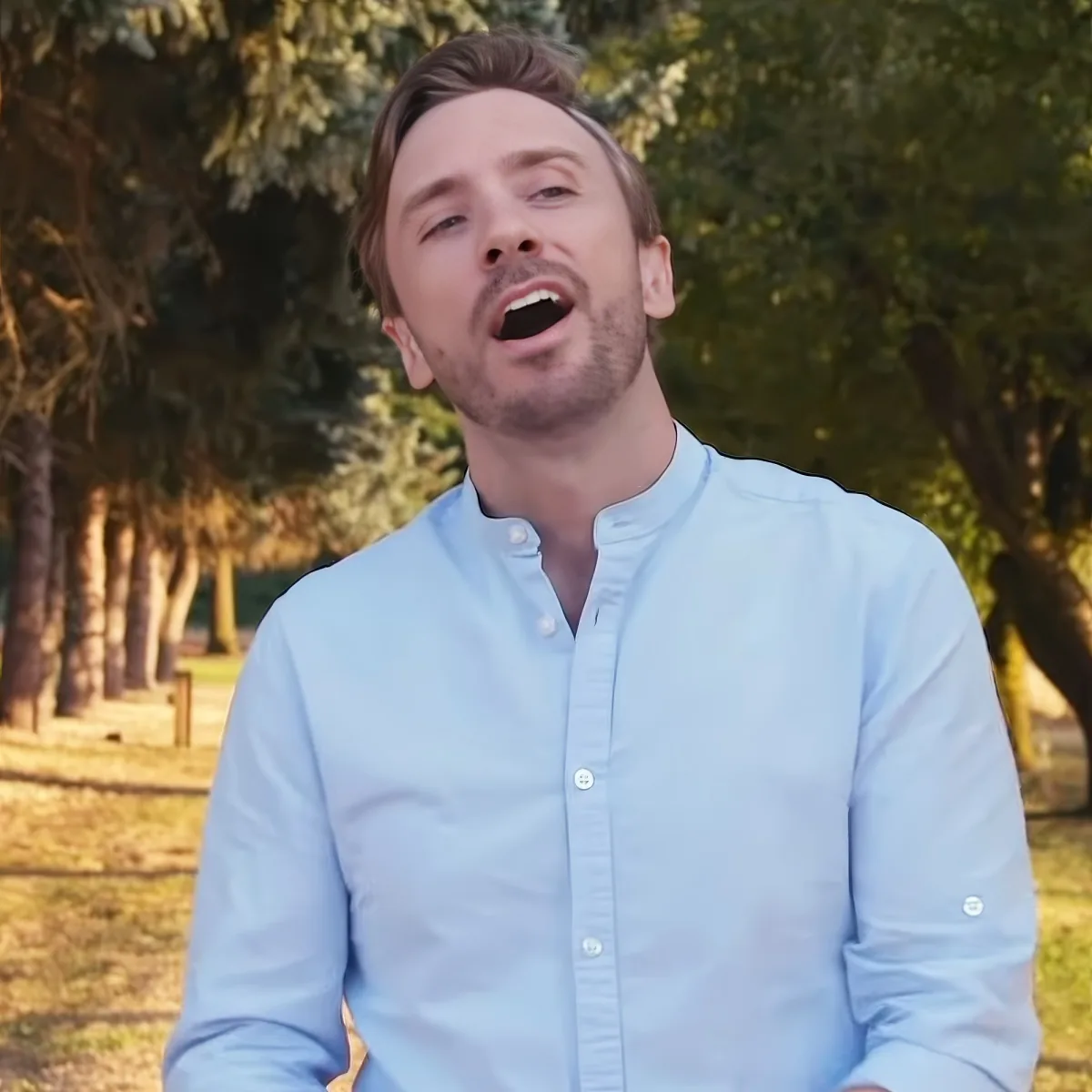 A man in a light blue shirt is sitting outdoors with trees in the background.