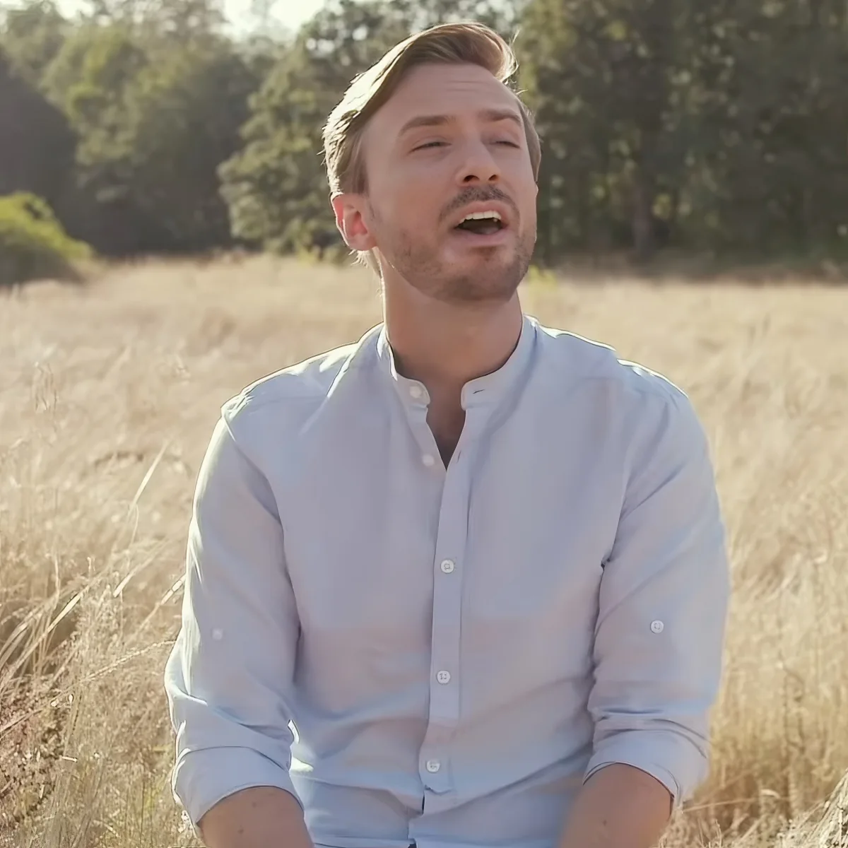 A man in a light blue shirt sits in a field of tall grass, eyes closed, mouth slightly open.