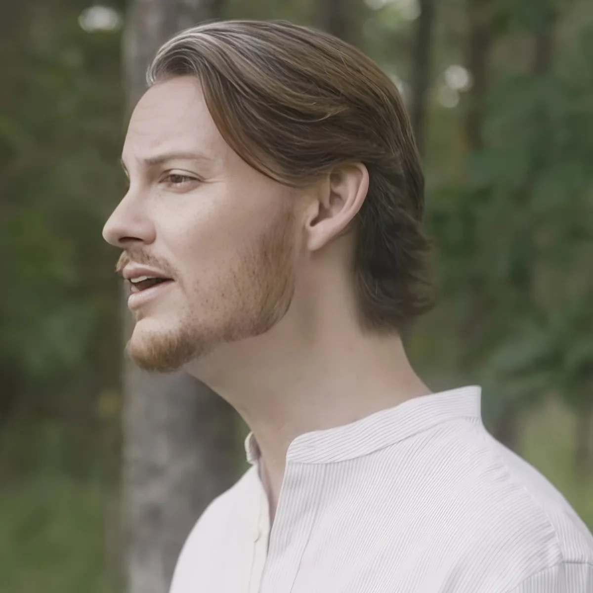 A man with long hair and a beard, wearing a white shirt, stands outdoors with trees in the background.