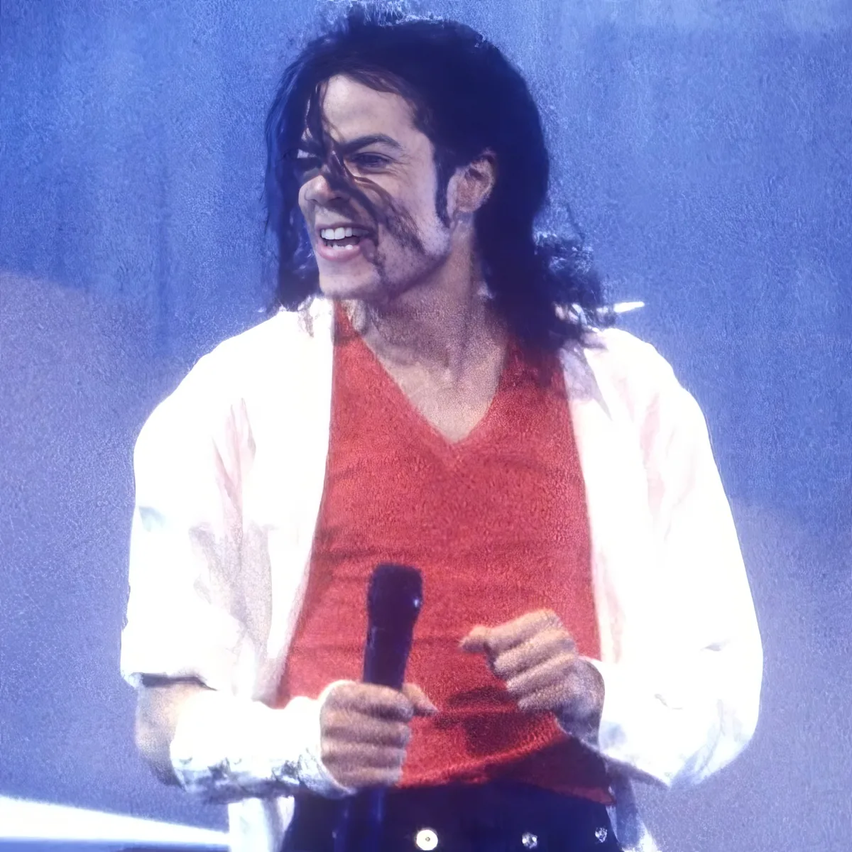 A person in a red shirt and white jacket holds a microphone, smiling, with long hair.