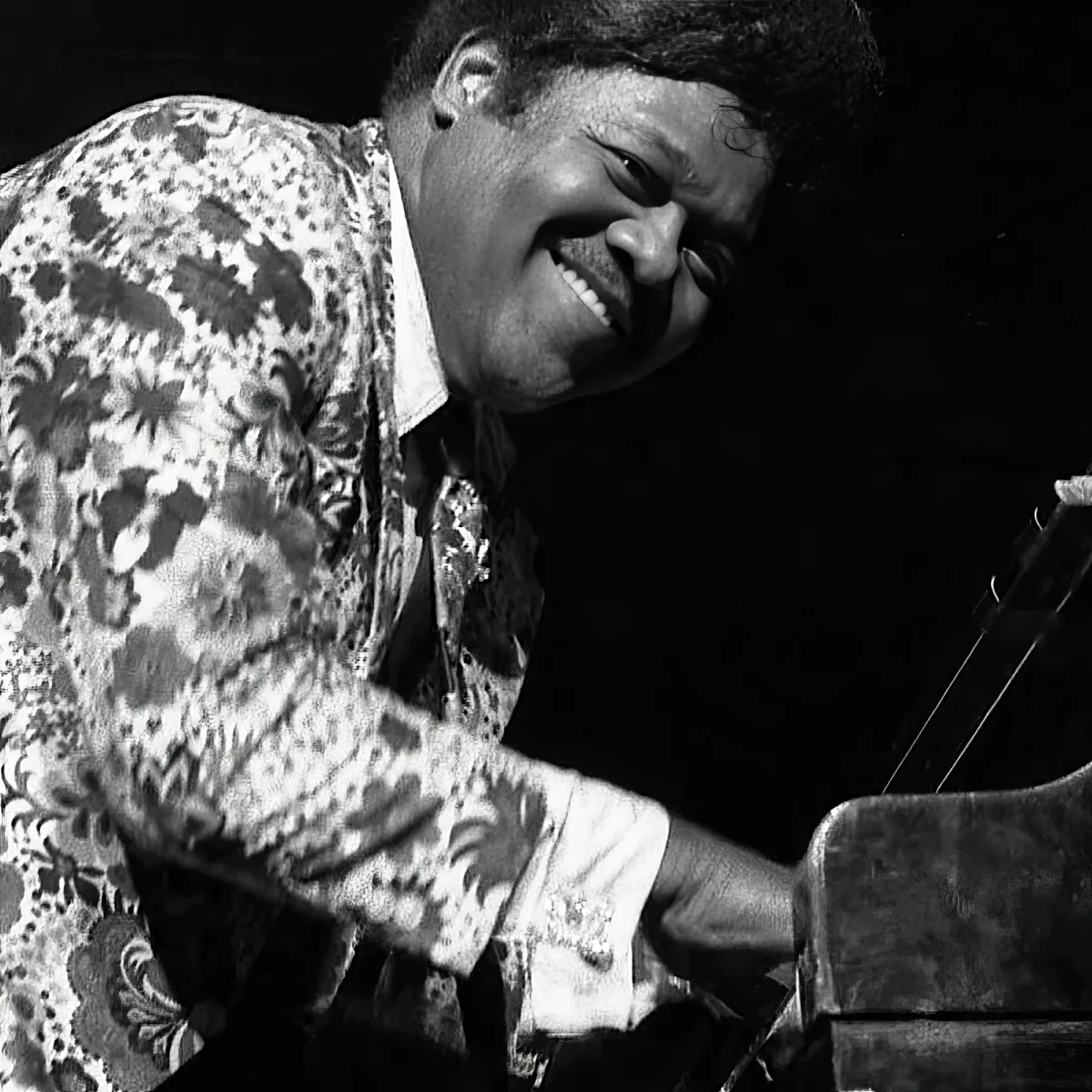 A smiling man in a patterned shirt plays a piano, captured in a black-and-white photograph.