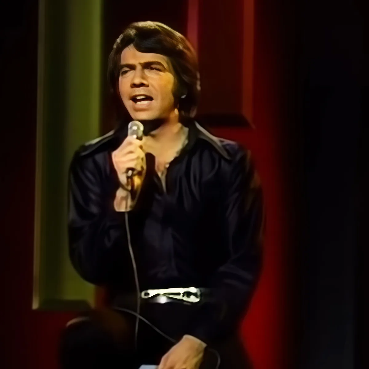 A man in a black jacket sings into a microphone on stage with red curtains.