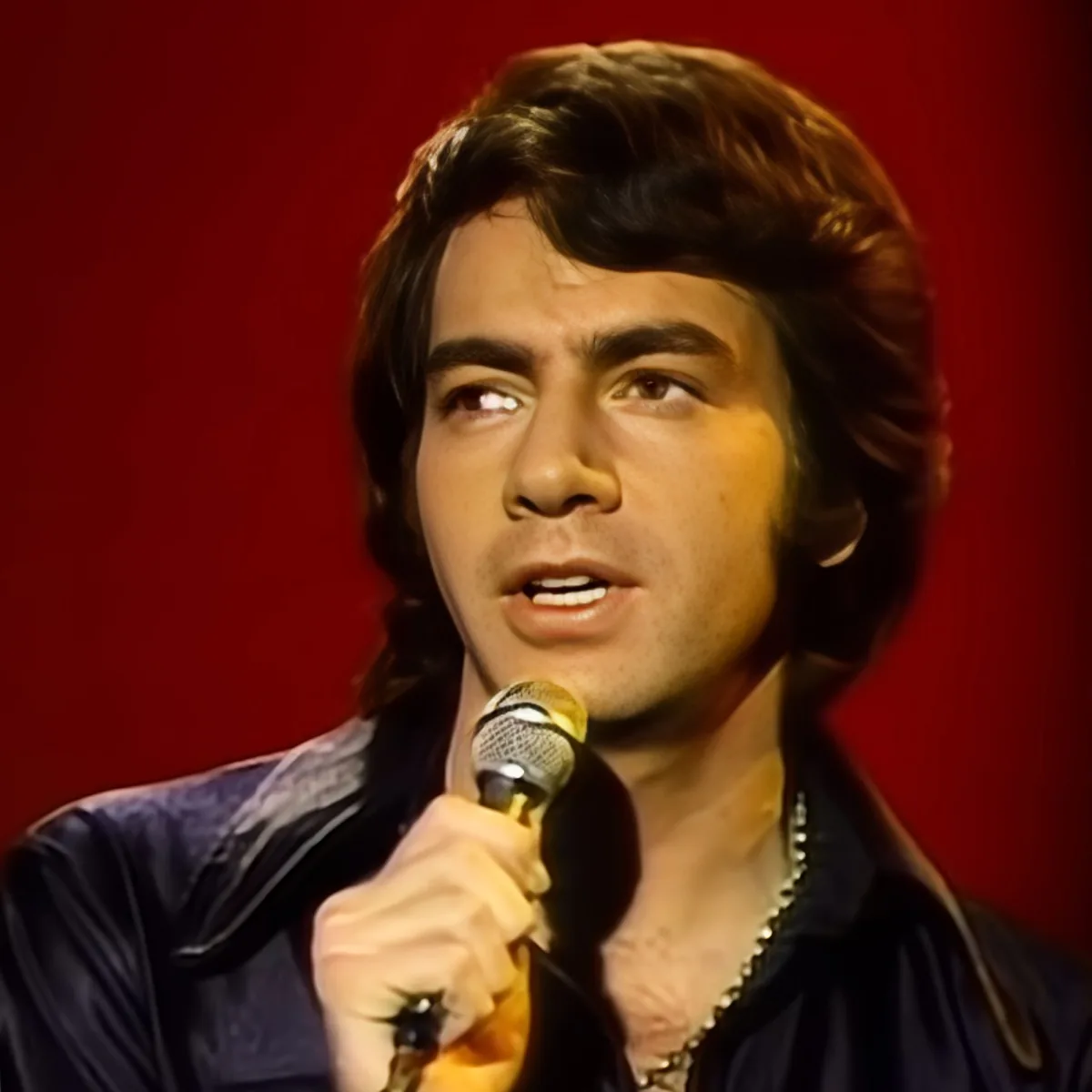 A man with dark hair and a gold chain holds a microphone, singing against a red background.