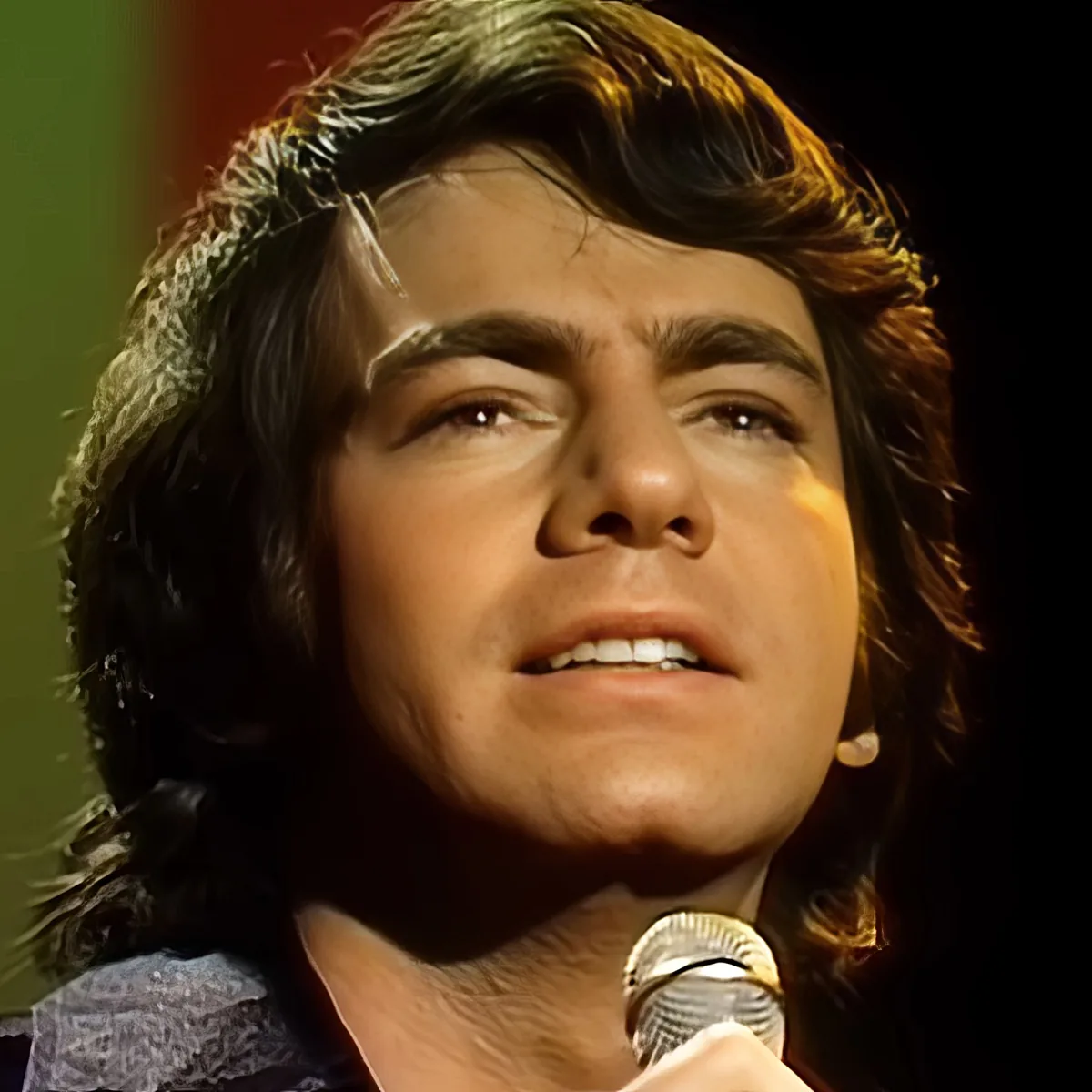 A person with long, wavy hair is singing into a microphone, wearing a patterned shirt.