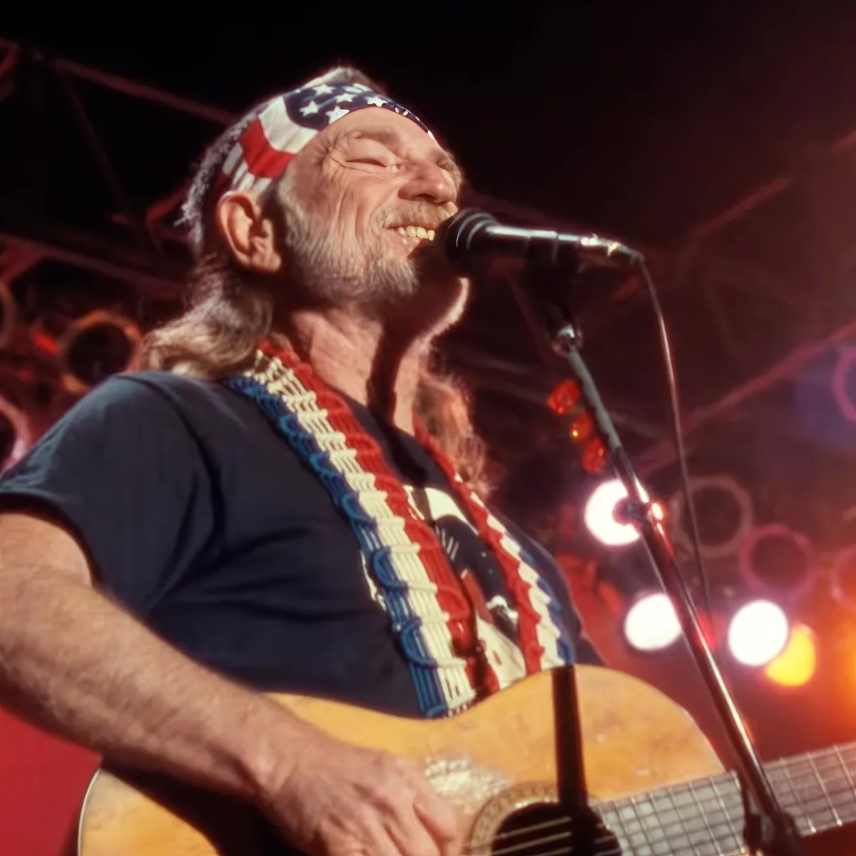 A man with a guitar and an American flag headband sings into a microphone on stage.