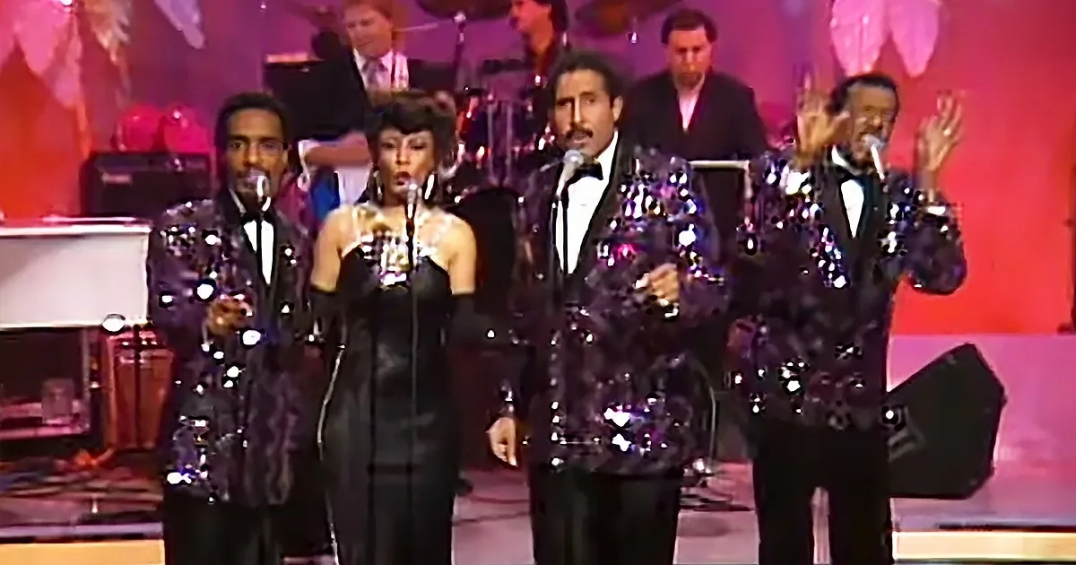 A group of performers in shiny, sequined outfits sing on stage with a band in the background.
