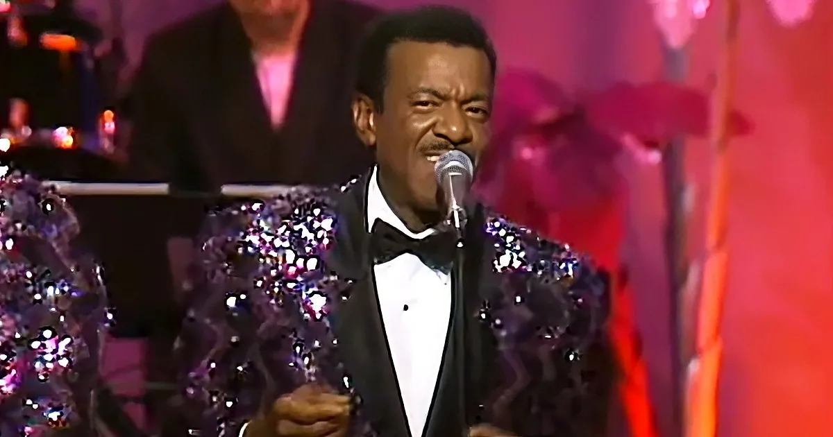 A man in a black sequined tuxedo sings into a microphone on stage.