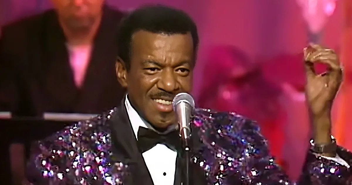 A man in a sparkling sequined suit sings into a microphone, smiling with a mustache.