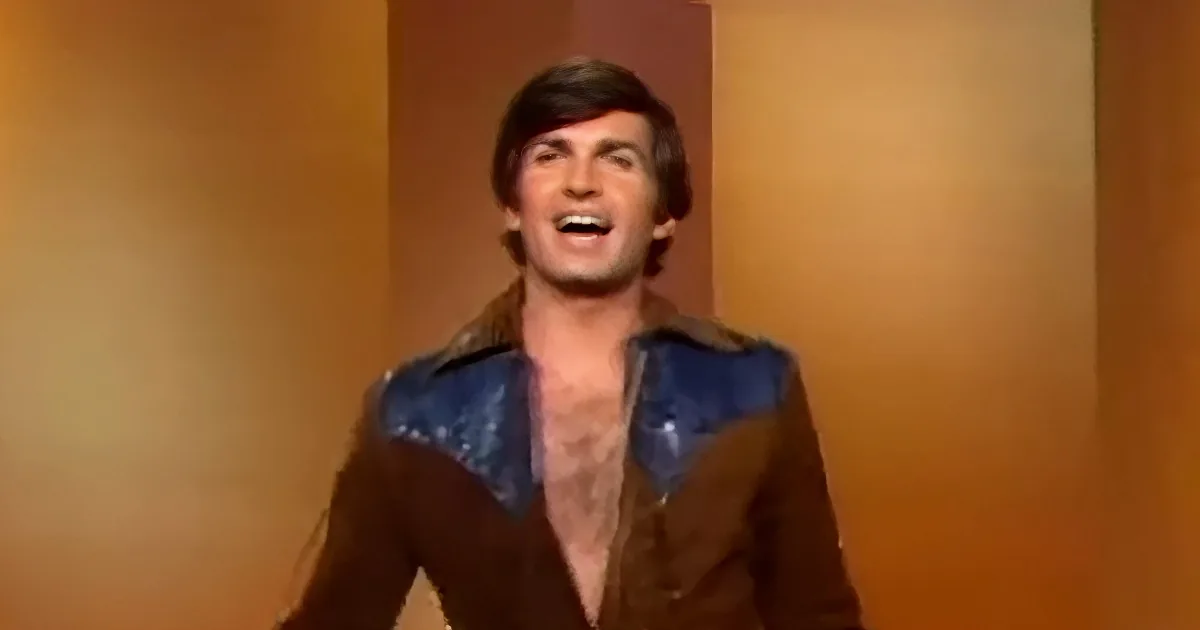 A man in a brown jacket with blue shoulder pads is smiling widely.