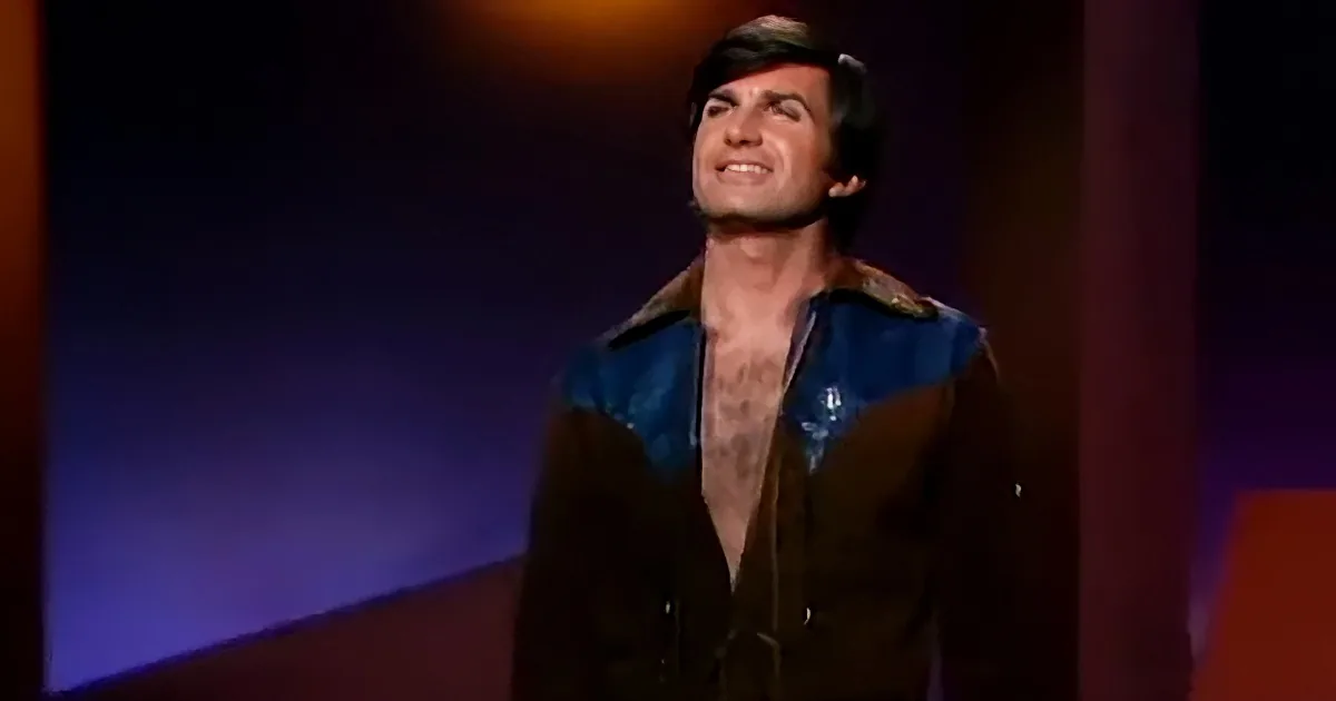 A man in a brown jacket with blue accents smiles upward on a stage with a dark background.