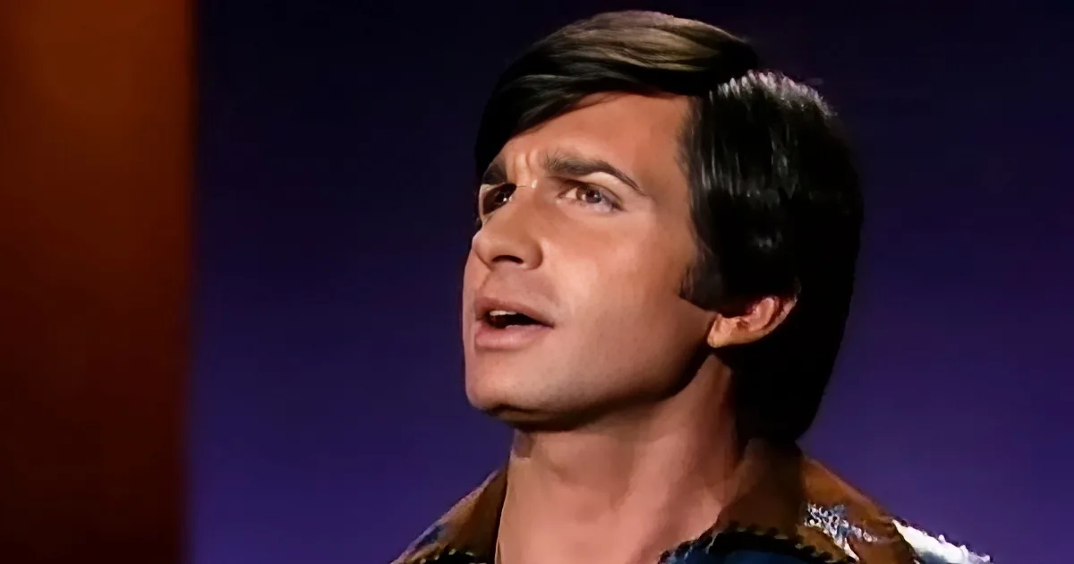 A man with dark hair and a patterned shirt is looking upwards with his mouth slightly open.