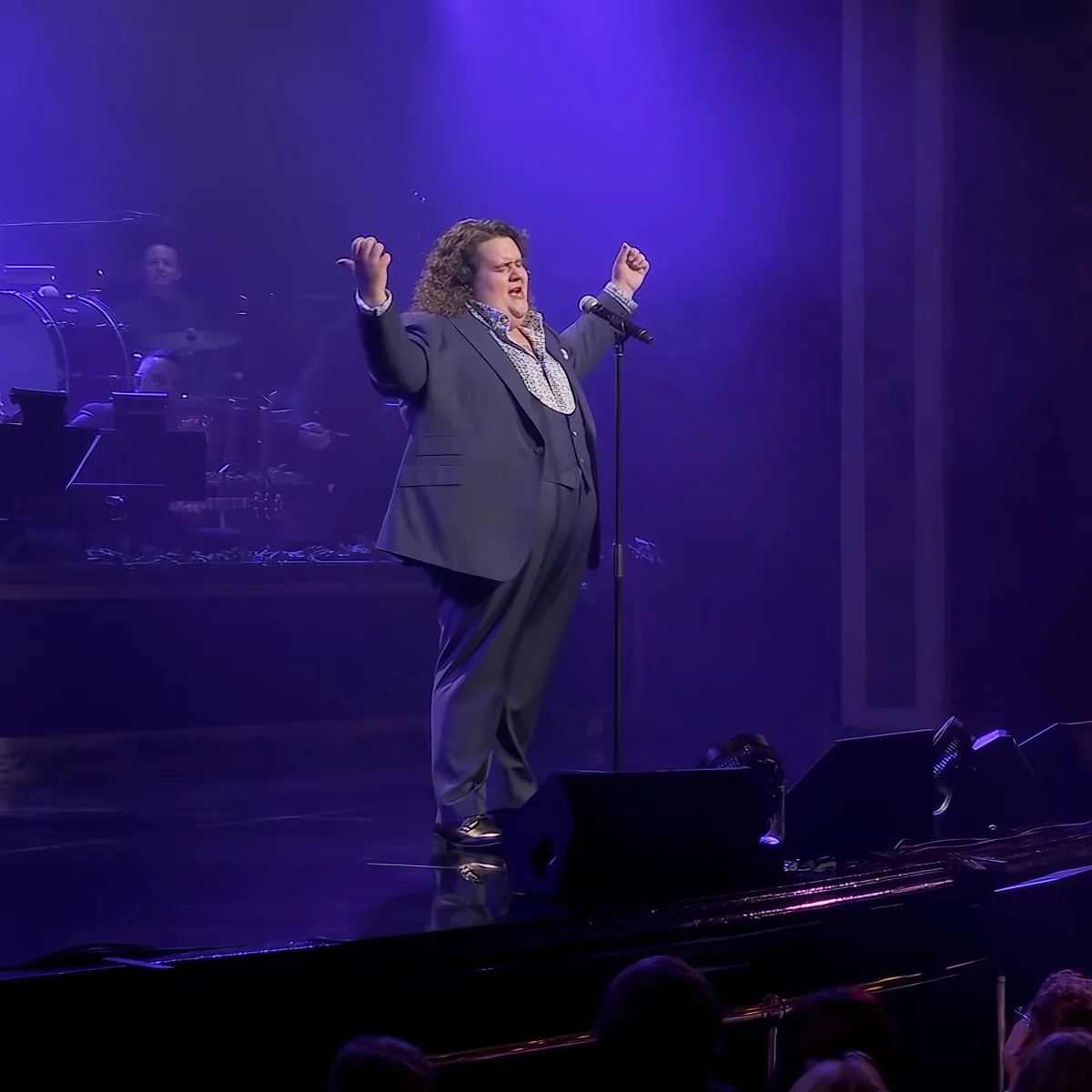 A person in a suit sings passionately on stage with a microphone, surrounded by a band and audience.