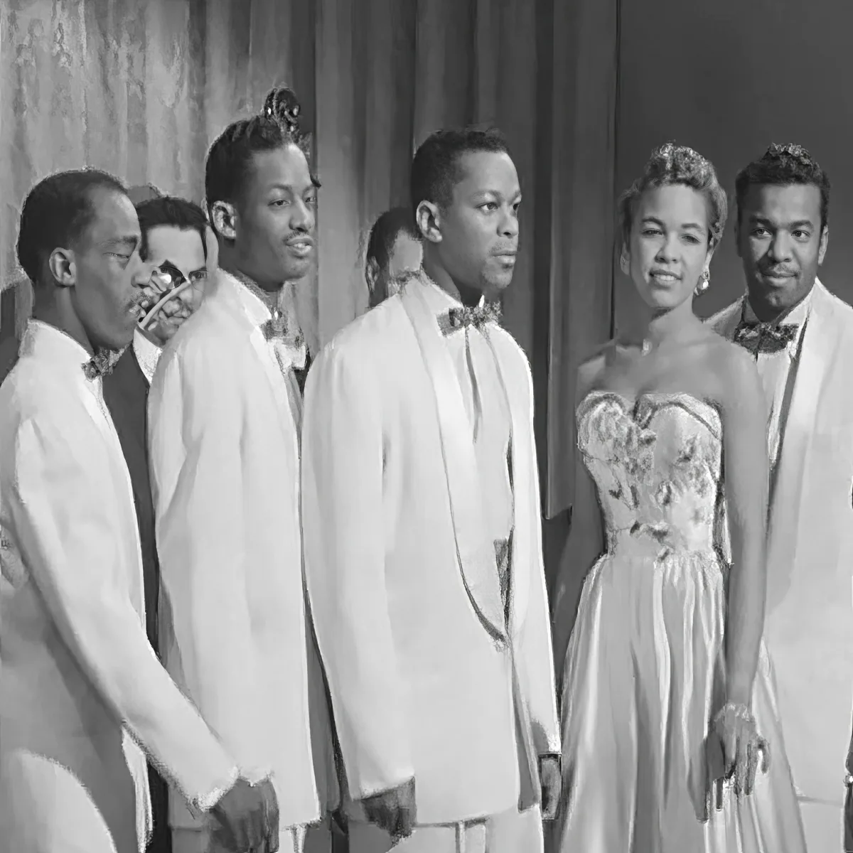 A group of elegantly dressed individuals, including a woman in a white dress and men in white suits, stand together.