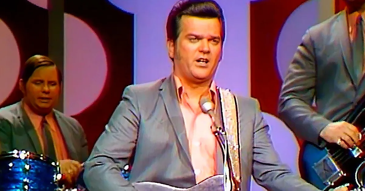 A man in a gray suit plays a banjo on stage with a drummer and another musician in the background.
