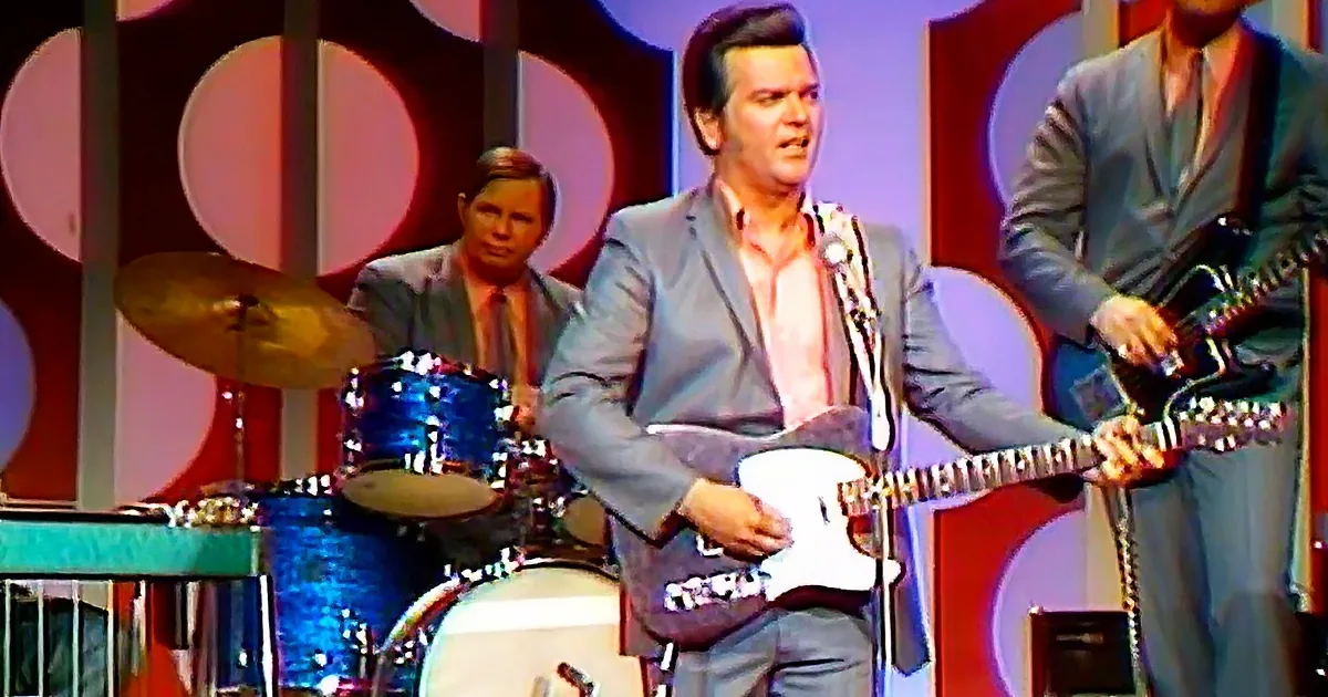 A man in a suit plays guitar on stage, with a drummer and another musician in the background.