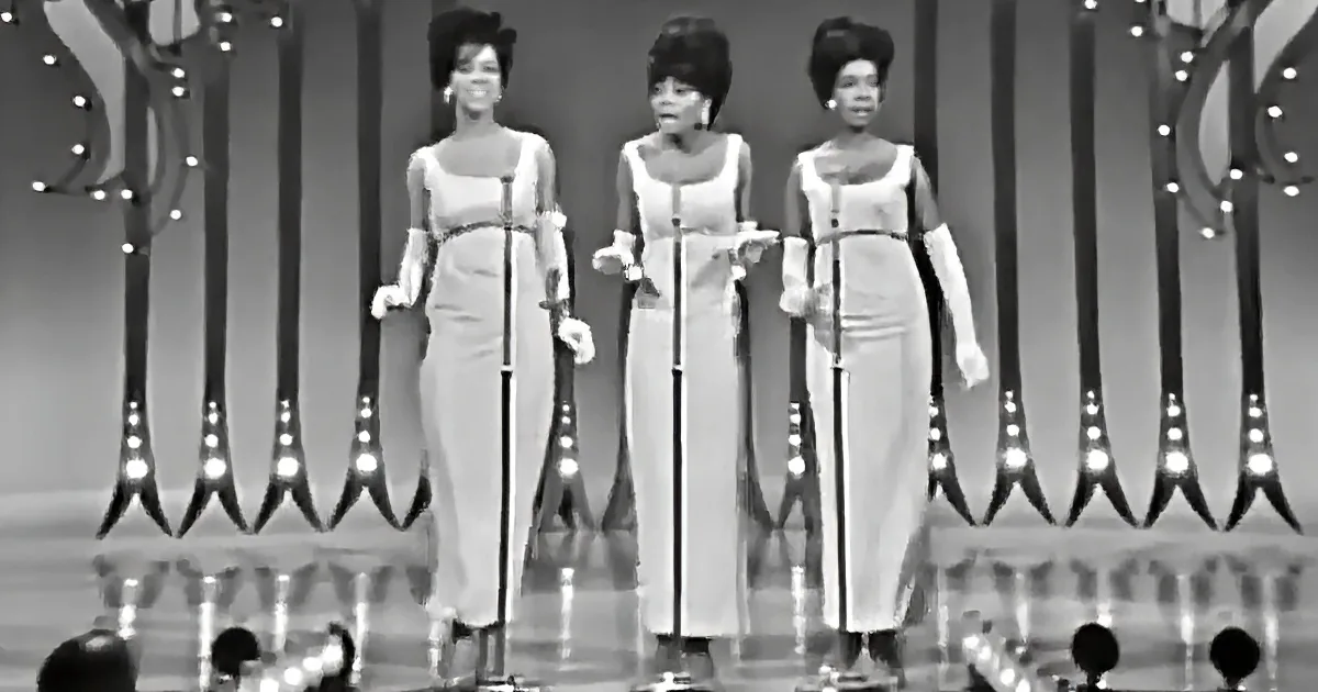 Three women in matching outfits sing on stage with microphones, surrounded by decorative lights.