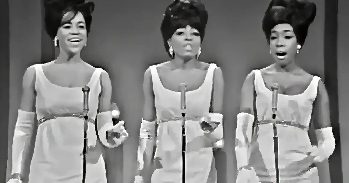 Three women in white dresses sing into microphones, wearing matching outfits and styled hair.